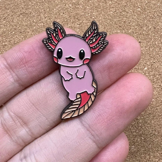 Pink axolotl-shaped enamel pin held between fingers on a brown background