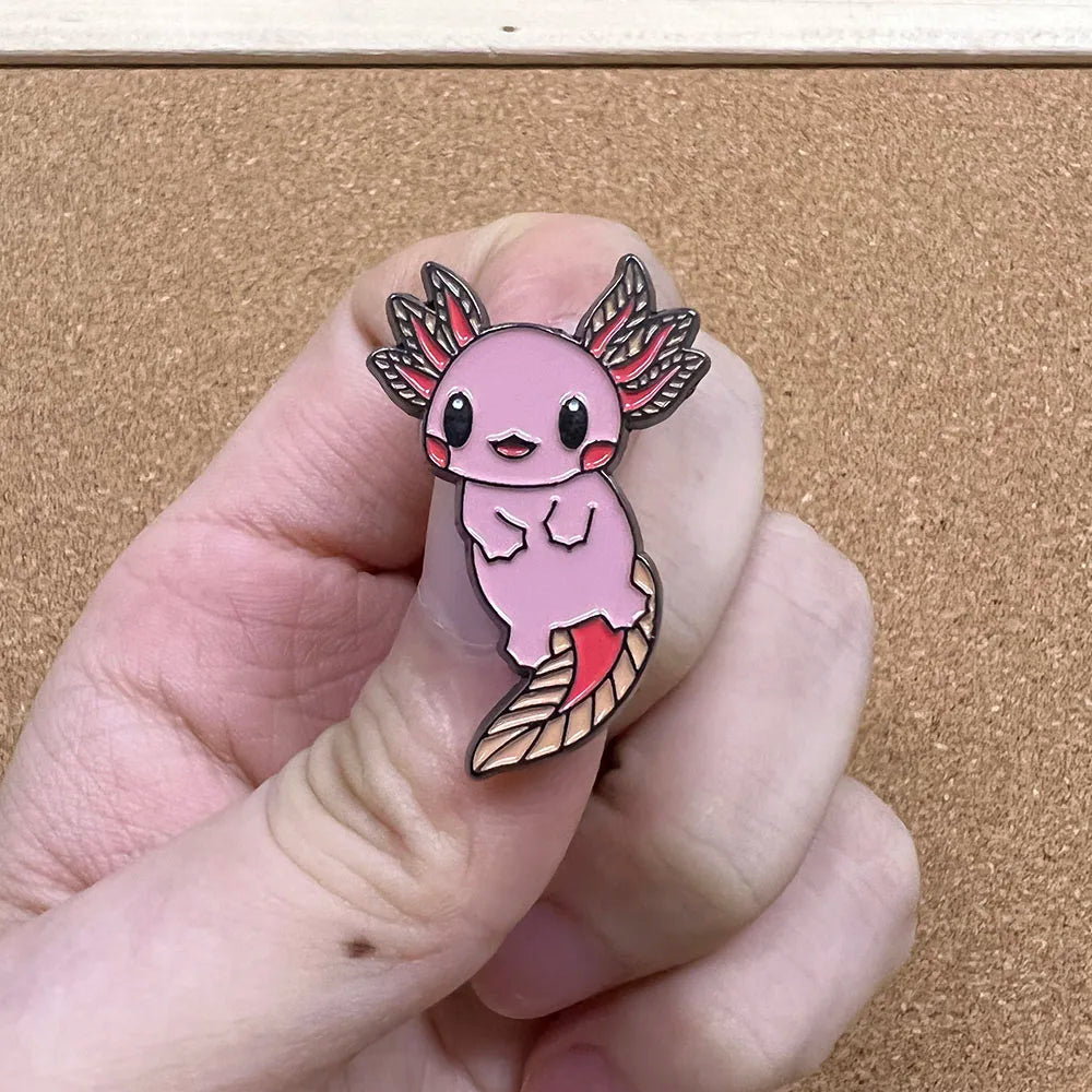 Pink axolotl enamel pin held between fingers against a beige background