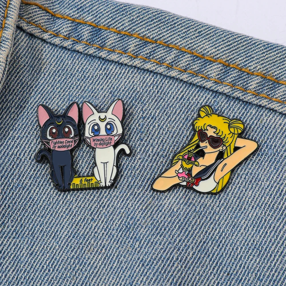 Two sailor moon character pins on a denim background, one featuring cartoon cats and the other a character with sunglasses.