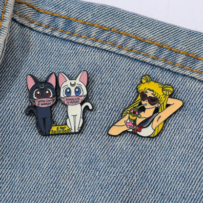 Two sailor moon character pins on a denim background, one featuring cartoon cats and the other a character with sunglasses.